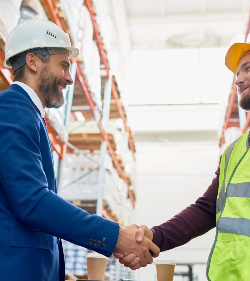 Waist up portrait of handsome mature businessman shaking hands with worker wearing hardhat standing against warehouse shelves in background
